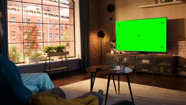 Young Couple Spending Time at Home, Sitting on a Couch and Watching TV with Green Screen Mock Up Display in Their Stylish Loft Apartment. Man and Woman Streaming Movie or Show and Eating Popcorn.