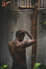 Shirtless muscular latin american male with tattoos standing taking shower outdoors after workout on the tropical leaves background. 