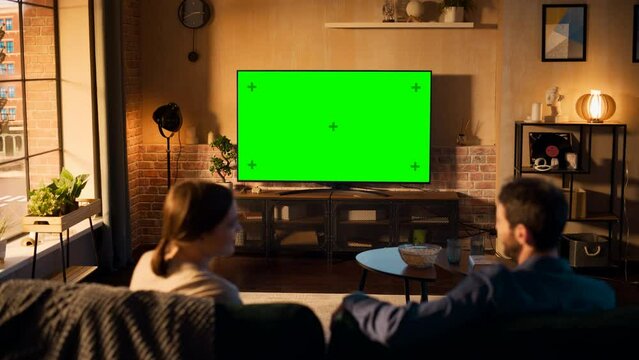 Young Couple Spending Time at Home, Sitting on a Couch and Watching TV with Green Screen Mock Up Display in Their Stylish Loft Apartment. Man and Woman Streaming Movie or Show. Shot From Back.