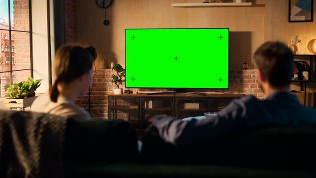 Authentic Couple Spending Time at Home, Sitting on a Couch and Watching TV with Green Screen Mock Up Display in Their Stylish Loft Apartment. Man and Woman Streaming Movie or Show. Shot From Back.