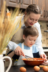 A young beautiful mother with her son in aprons at home in the kitchen prepare dough for baking cookies against the background of ears of wheat. On the table are eggs and a rolling pin.