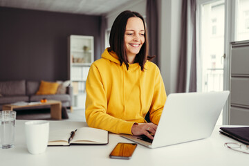 Young adult woman in yellow hoodie type on laptop. Remote education