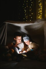 Two children with flashlight read a book under a blanket as a tent - obrazy, fototapety, plakaty