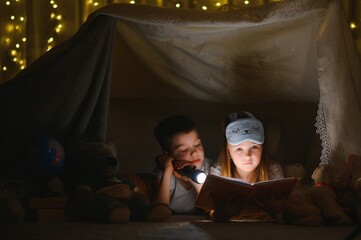 Fototapeta na wymiar Little kids involving in reading amazing book. They lying in nice toy tent in playroom. Boy holding flashlight in hand