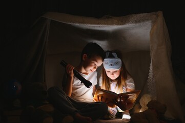 Siblings sit in a hut of chairs and blankets. Brother and sister reading book with flashlight at home - obrazy, fototapety, plakaty