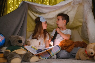 Obraz na płótnie Canvas Two children with flashlight read a book under a blanket as a tent