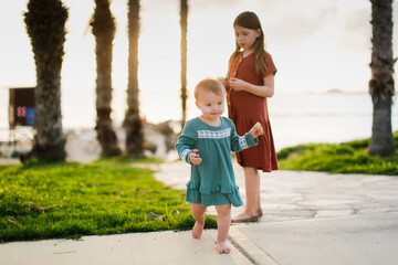 Cute European kid girl in dress on sea. Girl with her sister at sunset on ocean. Walks along seashore in silence at sunset. Kids run stone path near sea, happy childhood and sibling relationships