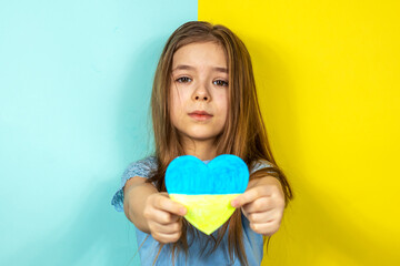 A girl in a blue jacket shows a heart painted in yellow-blue colors as a sign of love for Ukraine, close-up against the background of the Ukrainian flag. Stop the war in Ukraine. The concept of peace