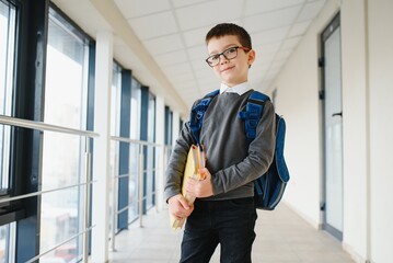Happy smiling boy in glasses is going to school for the first time. Child with school bag and book in his hand. Kid indoors of the class room . Back to school.