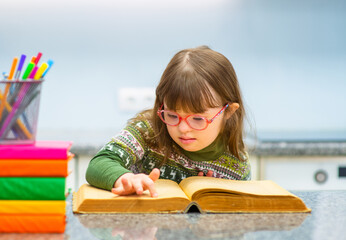 A girl with down syndrome with glasses sitting at a table and reading a big serious book swiping her finger across the page