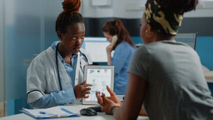 Obraz premium African american doctor and patient looking at human skeleton on tablet. Black people analyzing osteopathy figure of bones anatomy on display for orthopedic diagnosis and healthcare.