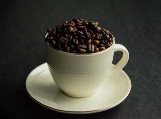 Brązowe ziarenka kawy wsypane do białej filiżanki z uszkiem i złotą otoczką leżącej na spodku na czarnym tle.