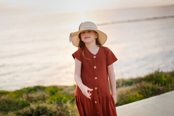 Cute long-haired European baby girl in a dress with a hat on the sea coast. Walking along the seashore in silence at sunset. Beautiful sky in a hat and wind