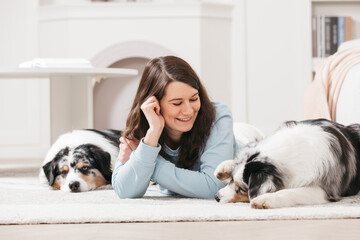 A young woman and two dogs are lying on the floor in a room. They are happy, laughing and playing. Aussie breed.