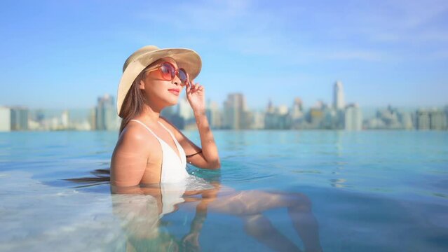 Classy asian woman in rooftop swimming pool above modern cityscape skyline enjoying on afternoon sun