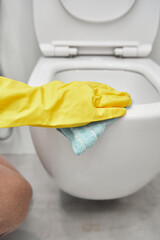A woman's hand in yellow rubber gloves wipes the toilet bowl with a rag.