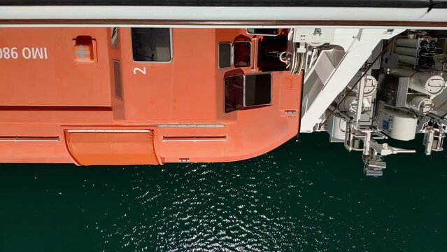 Big safety boat hanging from the side of a cruiser seen from above. Someone from maintenance is inside and opens the different windows on the boat top to carry a routine check.