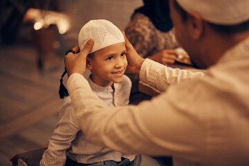 Happy Muslim boy looks at his father who is adjusting him traditional skullcap at home.