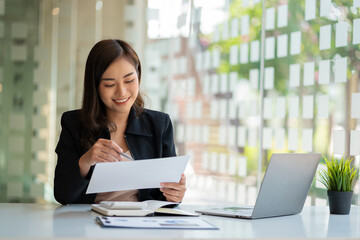 Asian businesswoman working in financial accounting analyzes a chart using a laptop calculator with documents and holding a pen sitting on a chair at an office desk.