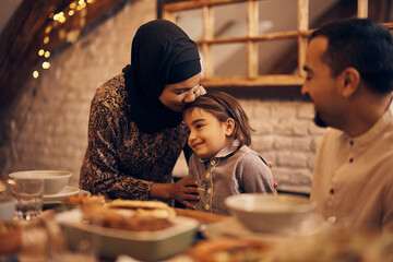 Loving Muslim mother kisses her daughter during family meal at dining table.