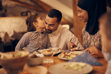 Loving Muslim girl kisses her father during family meal at dining table.