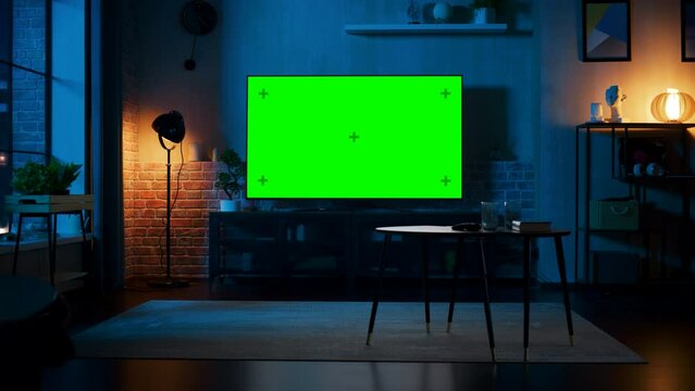 Stylish Loft Apartment Interior with TV Set with Green Screen Mock Up Display Standing on Television Stand. Empty Living Room at Home with Chroma Key Placeholder on Monitor. Zoom In Night Shot.