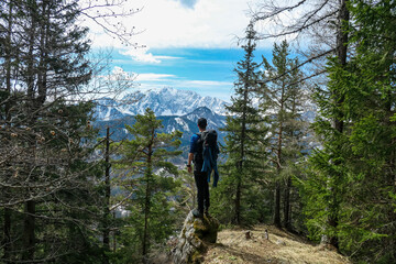 Fototapeta premium Man with backpack standing on a rock with scenic view of snow capped mountain peaks of Karawanks near Sinacher Gupf in Carinthia, Austria. Mount Wertatscha is visible through dense forest in spring