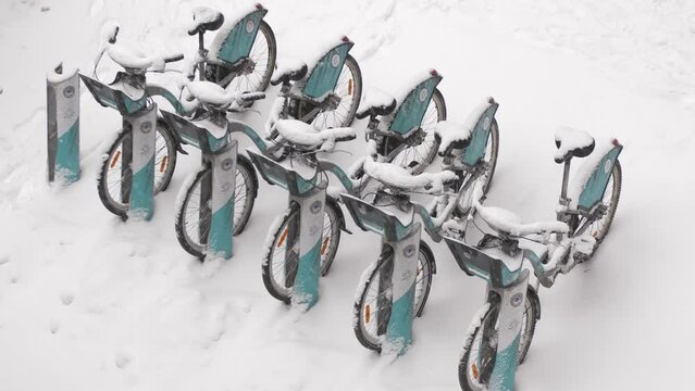 Bicycles covered with snow stand during a snowfall at a bike sharing station.