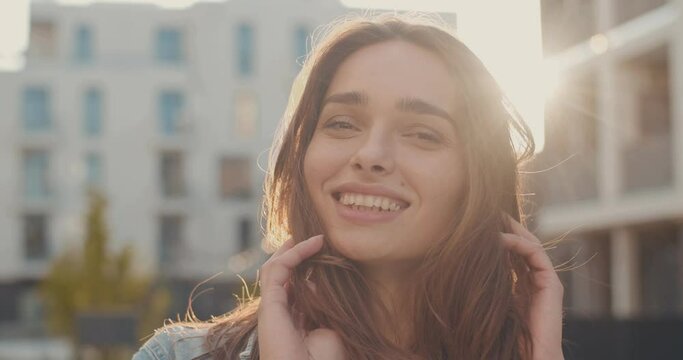 Portrait of the young Caucasian styish and beautiful girl smiling cheerfully to the camera and stiring up her hair while posing to the camera as professional model at the street. Close up. Outdoor.