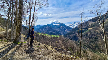 Man with backpack and scenic view of snow capped mountain peaks of Karawanks near Sinacher Gupf in Carinthia, Austria. Mount Hochstuhl (Stol) visible through forest in spring. Rosental sunny day. Awe