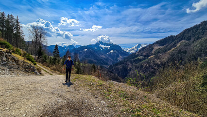 Fototapeta na wymiar Man with backpack and scenic view of snow capped mountain peaks of Karawanks near Sinacher Gupf in Carinthia, Austria. Mount Hochstuhl (Stol) visible through forest in spring. Rosental sunny day. Awe