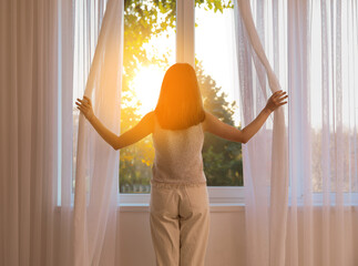 Pretty young woman opening curtains in room on sunny day, back view