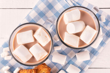 Two mugs with hot cocoa drink and marshmallow on a blue cloth napkin on a white wooden background....
