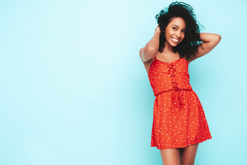 Portrait of beautiful black woman with afro curls hairstyle. Smiling model dressed in red summer dress. Sexy carefree female posing near blue wall in studio. Tanned and cheerful