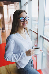 A young woman in glasses of European appearance, she is in a white shirt drinking coffee and looking out the window