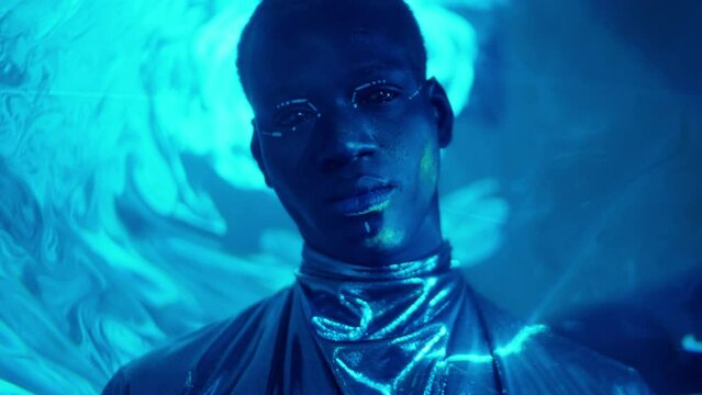 Creative portrait of futuristic African American man with tribal makeup wearing silver outfit posing on camera in studio filled with blue laser light
