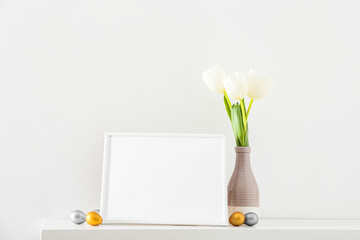 Blank photo frame, Easter eggs and vase with tulips on table near light wall