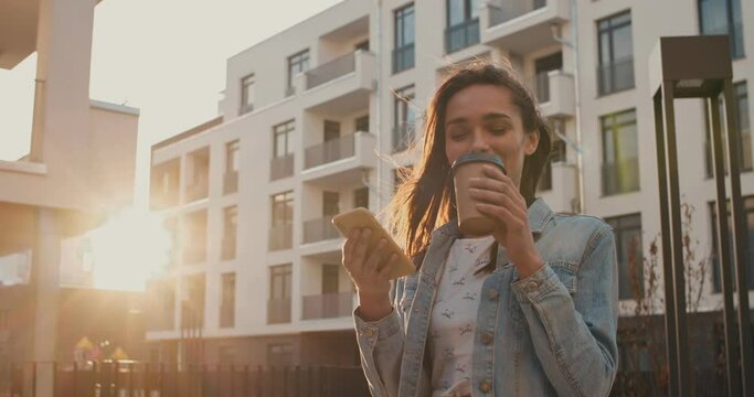 Caucasian smiled young attractive woman using her smartphone for social media visit and sipping coffee to-go while standing outside block of apartments in the sleeping area neighborhood.