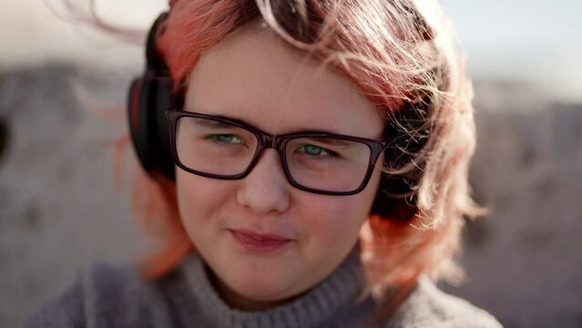 pretty preteen girl with glasses and modern headphones for listening to music is sitting on street