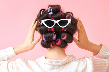 Beautiful young woman in pajamas, hair curlers and with sunglasses on pink background, back view