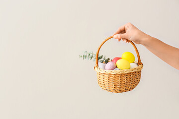 Woman holding gift basket with painted Easter eggs and cookies on light background