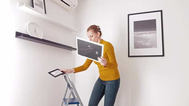 Woman on the ladder hanging pictures and photos on the shelf and wall at home.