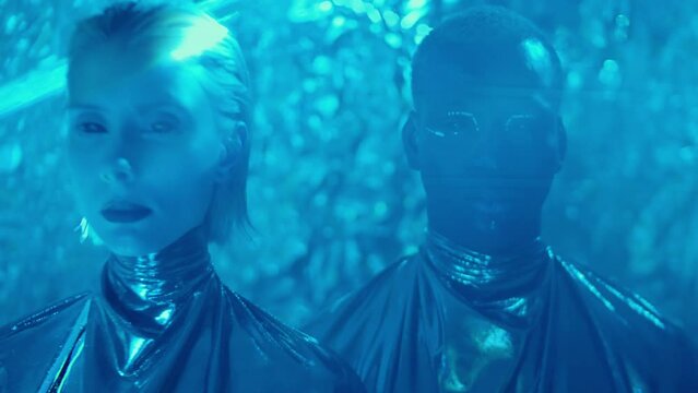 Studio shot of alien woman and man with creative makeup and blackout lenses wearing silver outfits posing for camera in blue laser light