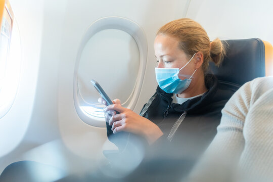 A Young Woman Wearing Face Mask, Using Smart Phone While Traveling On Airplane. New Normal Travel After Covid-19 Pandemic Concept.