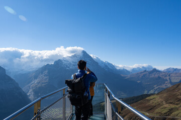 Fototapeta na wymiar The back of Asian male traveler with backpack standing at the end of the steel walkway in mountain cliff, taking photos of the amazing Matterhorn mountain peak covered with clouds in a clear blue sky.