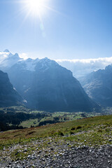 A vertical image, top view of a beautiful mountain valley where the mountain peak touches the clouds, with rural villages on green meadow hills with pine trees below, in a clear blue sky with sunshine © Siam Stock