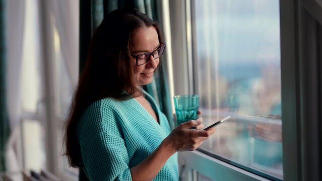 pretty middle-aged woman is sending message by apple in smartphone and drinking juice near window