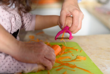 Fototapeta na wymiar Little cute baby toddler girl with grandmother in the kitchen peeling carrots with carrot peeler on chopping board. Child help at home, closeup of hands.Cooking with kids, healthy food, family love.