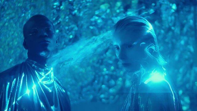 Mesmerized extraterrestrial woman and man with fantasy makeup and dark eyes wearing metallic gowns posing in fog and blue laser light in studio
