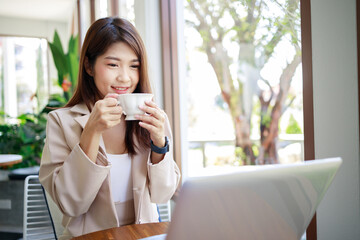 attractive Asian businesswoman drinking coffee and working by laptop in the coffee shop. Confident and smart woman and business concept.